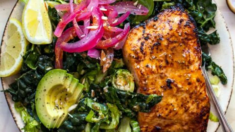 A plate of greens topped with sliced avocado, pickled red onion, and salmon fillet.