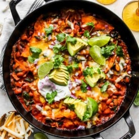 Skillet of vegetable enchiladas topped with avocado slices, cilantro, and lime wedges.