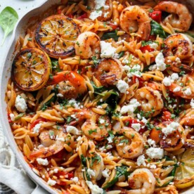 Shrimp and orzo in a white skillet topped with lemon wedges and feta