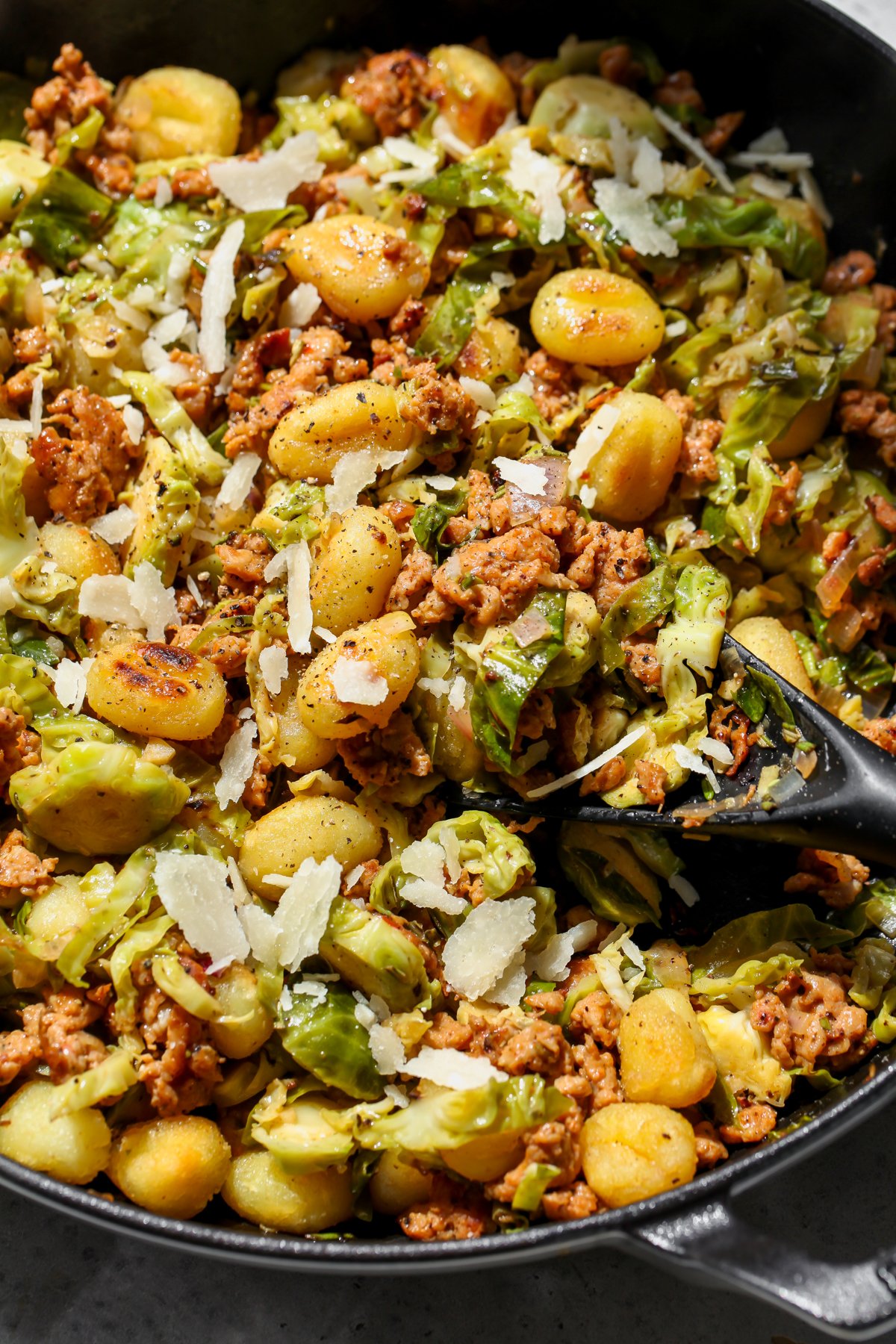 A large spoon scooping up a portion of gnocchi, sausage, and Brussels sprouts in a pan.