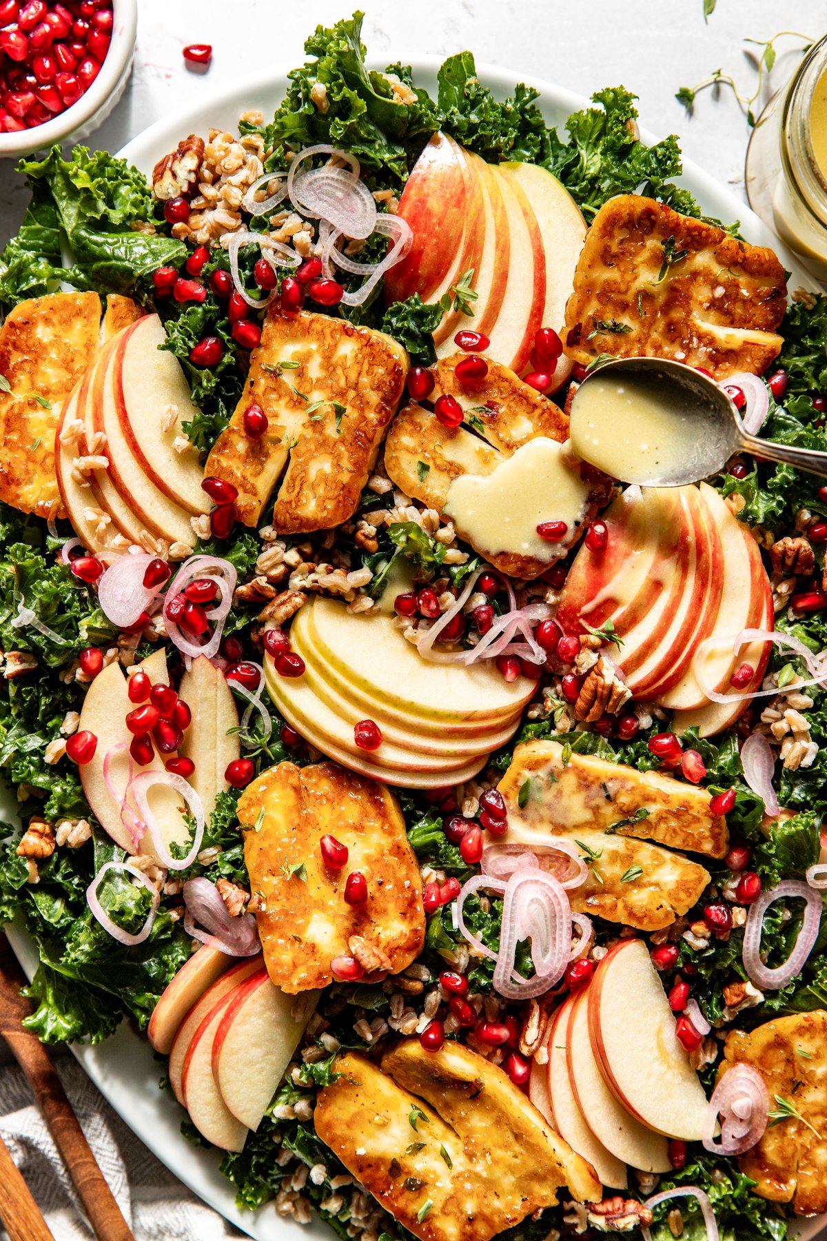 A spoon drizzling cider dressing over halloumi salad on a large white platter.
