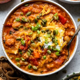 A bowl of turkey pumpkin chili topped with pepitas, cheese, and green onion styled on a gray surface.