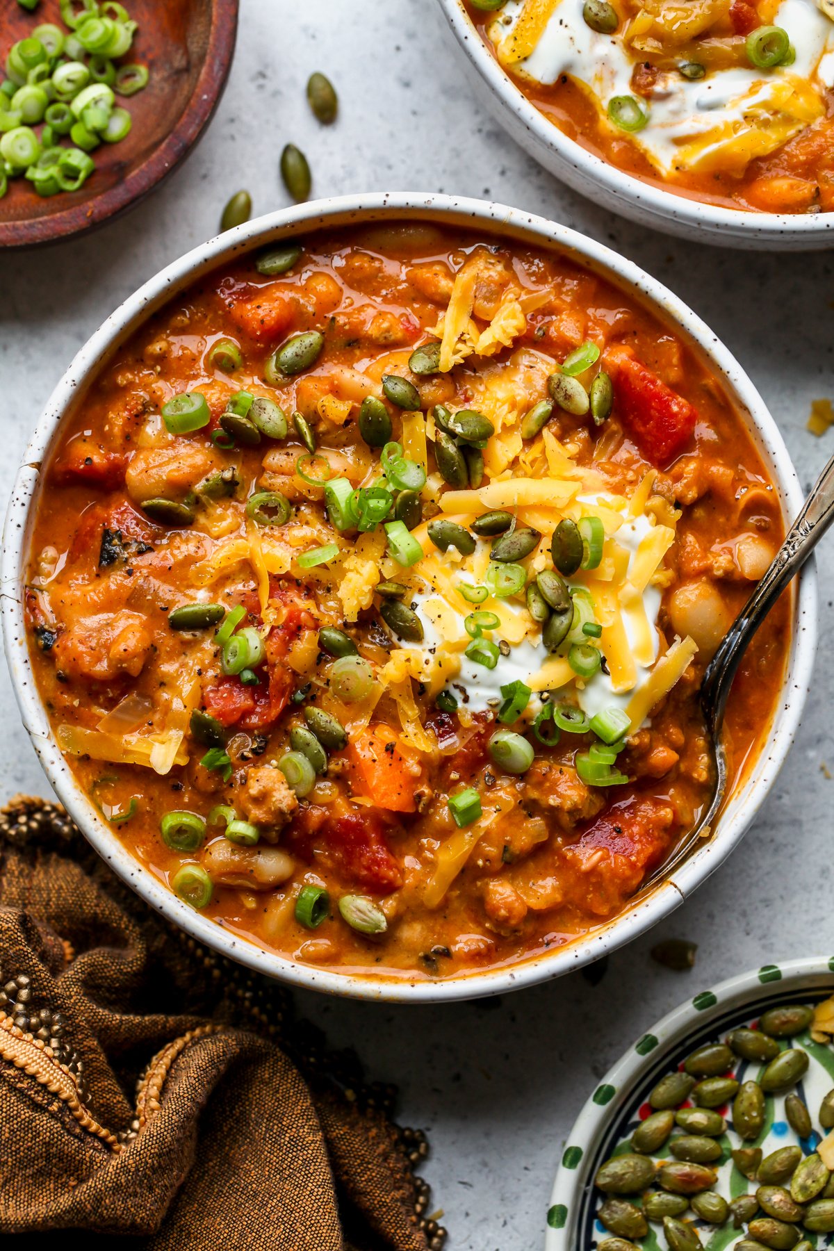 A bowl of turkey pumpkin chili topped with pepitas, cheese, and green onion styled on a gray surface.