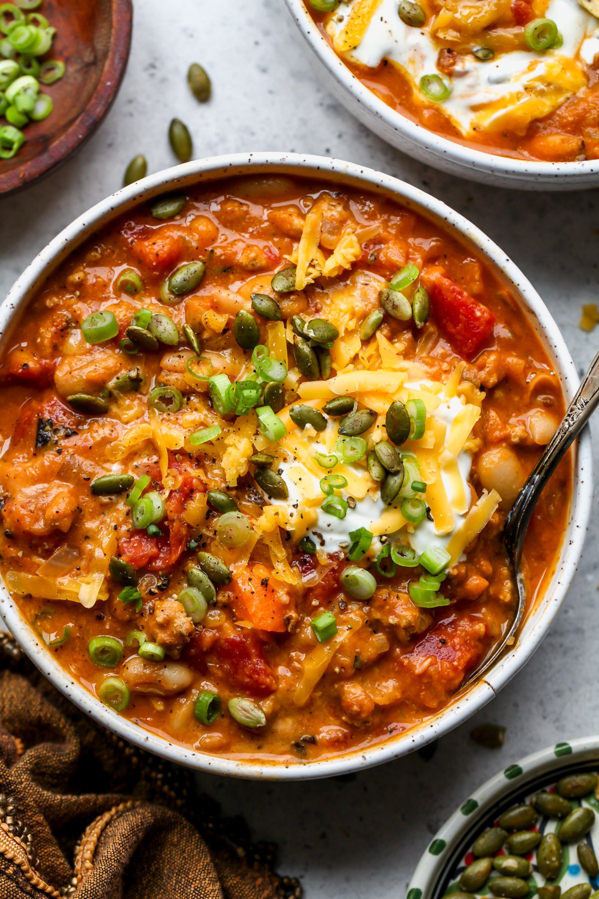 A tan bowl filled with a serving of turkey pumpkin chili topped with sour cream, shredded cheese, pepitas, and green onion.