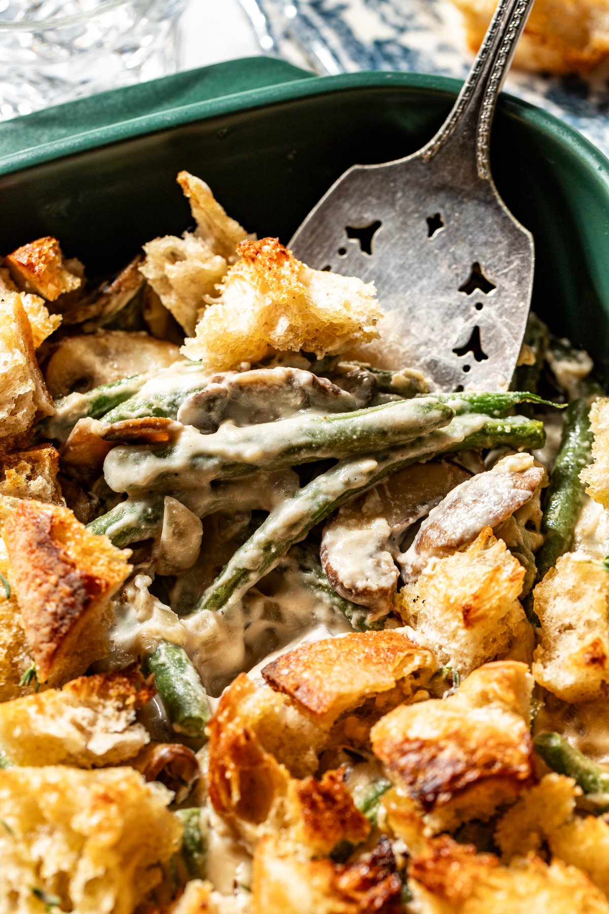 A spatula digging into a dish of green bean casserole topped with sourdough croutons.