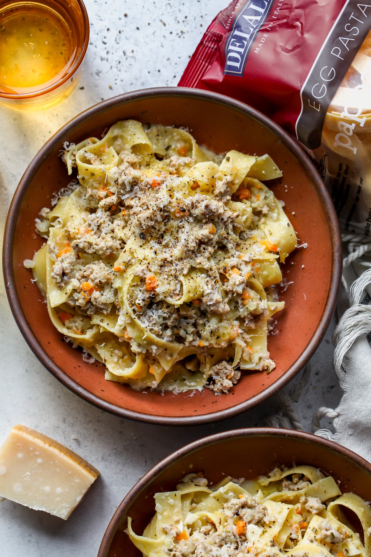 White bolognese with pappardelle pasta styled in two maroon bowls with a bag of pasta resting on the right side.