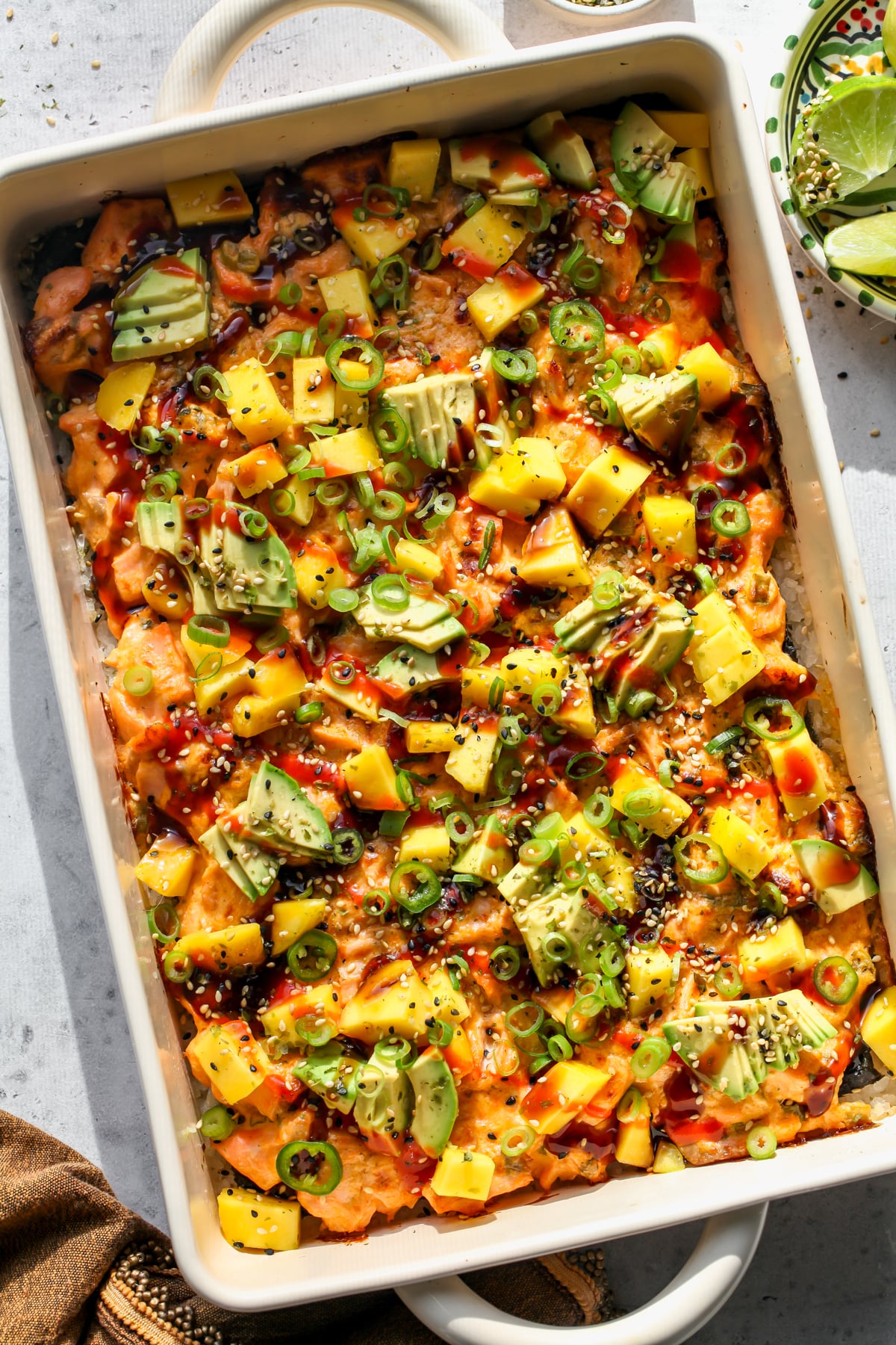 Salmon sushi bake styled in a rectangular casserole dish topped with avocado, mango, scallions, and sriracha.