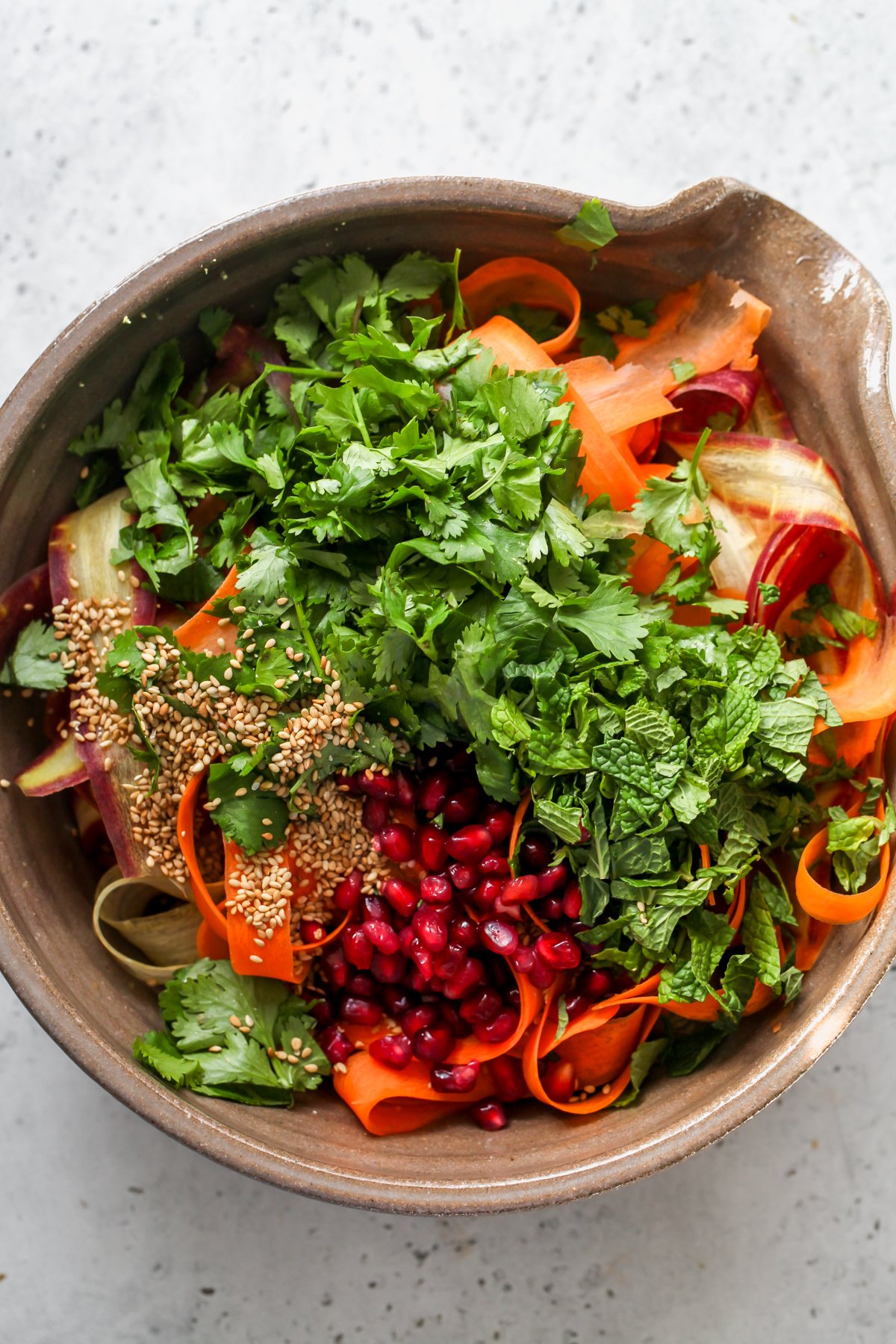 Cilantro, mint, pomegranate seeds, and sesame seeds being added to the bowl of carrots and dressing.