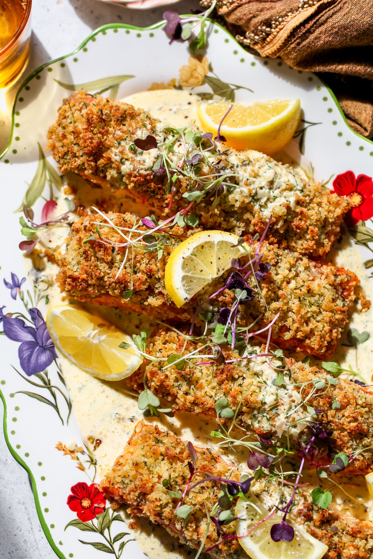 Panko Crusted Salmon with Creamy Dill Sauce styled on a floral platter topped with lemon slices and fresh herbs.