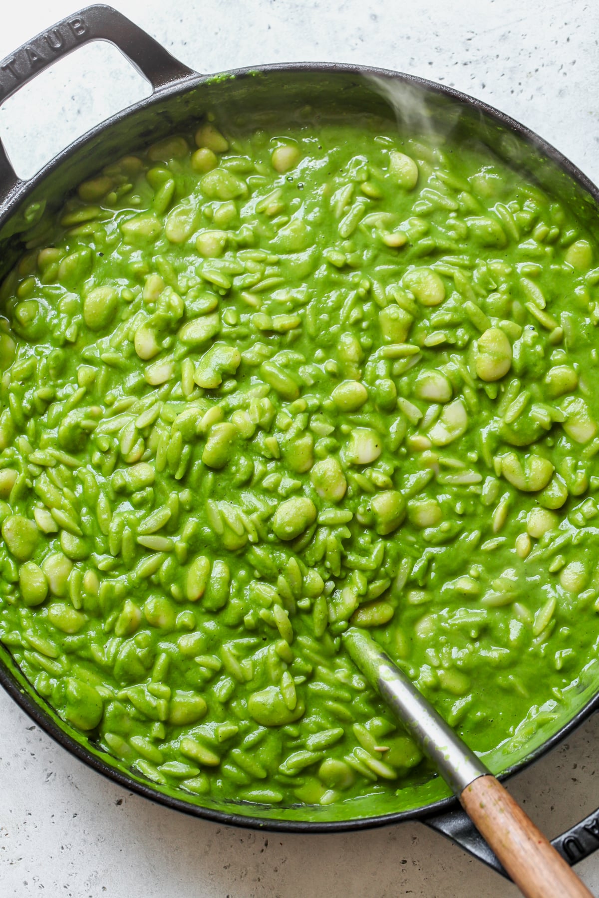 Beans, orzo, and green sauce being simmered and stirred in a large skillet.