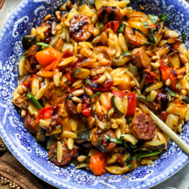 Chicken sausage orzo with burst tomatoes in a blue and white floral pasta bowl being stirred with a gold spoon.