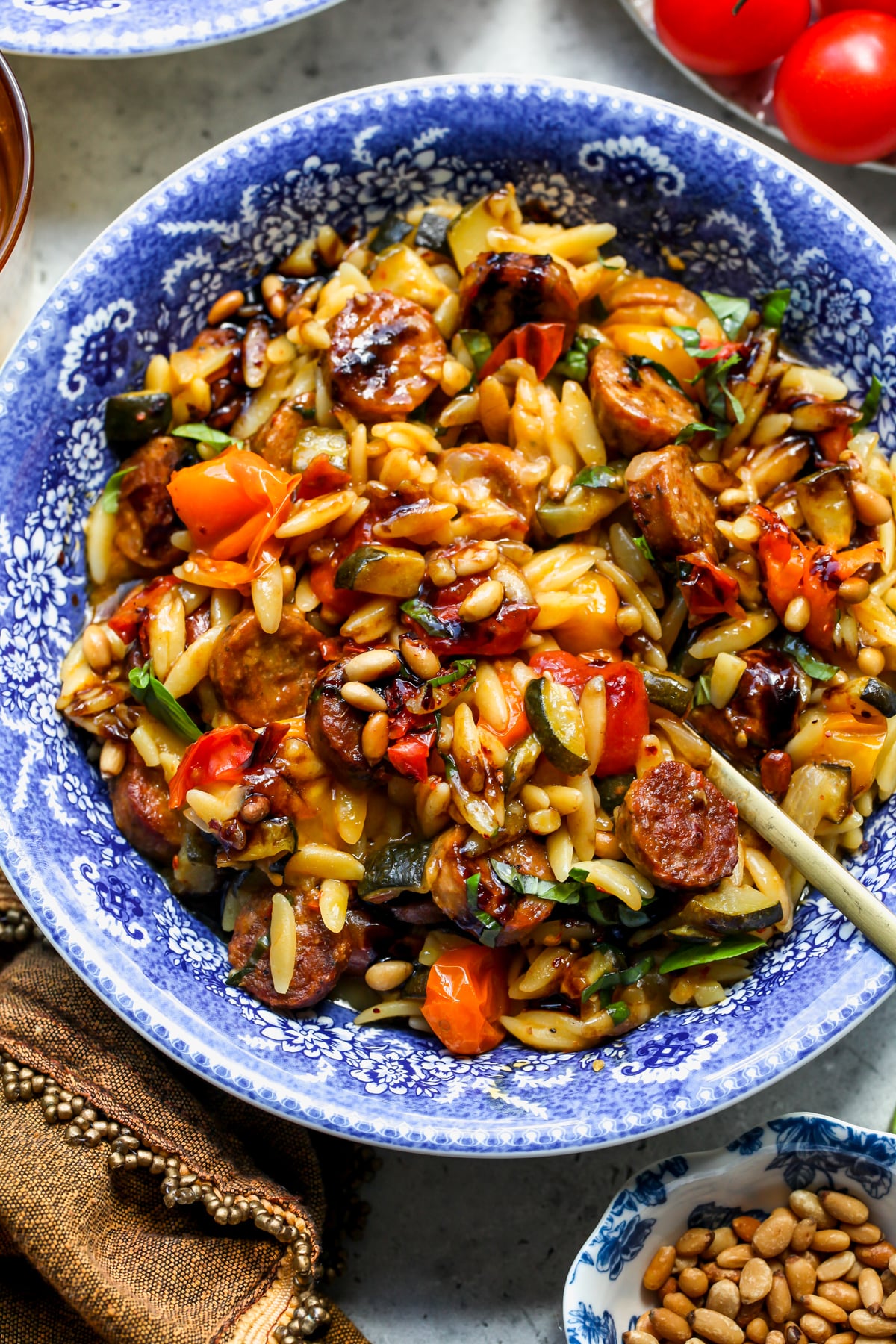 Chicken sausage orzo with burst tomatoes in a blue and white floral pasta bowl being stirred with a gold spoon.