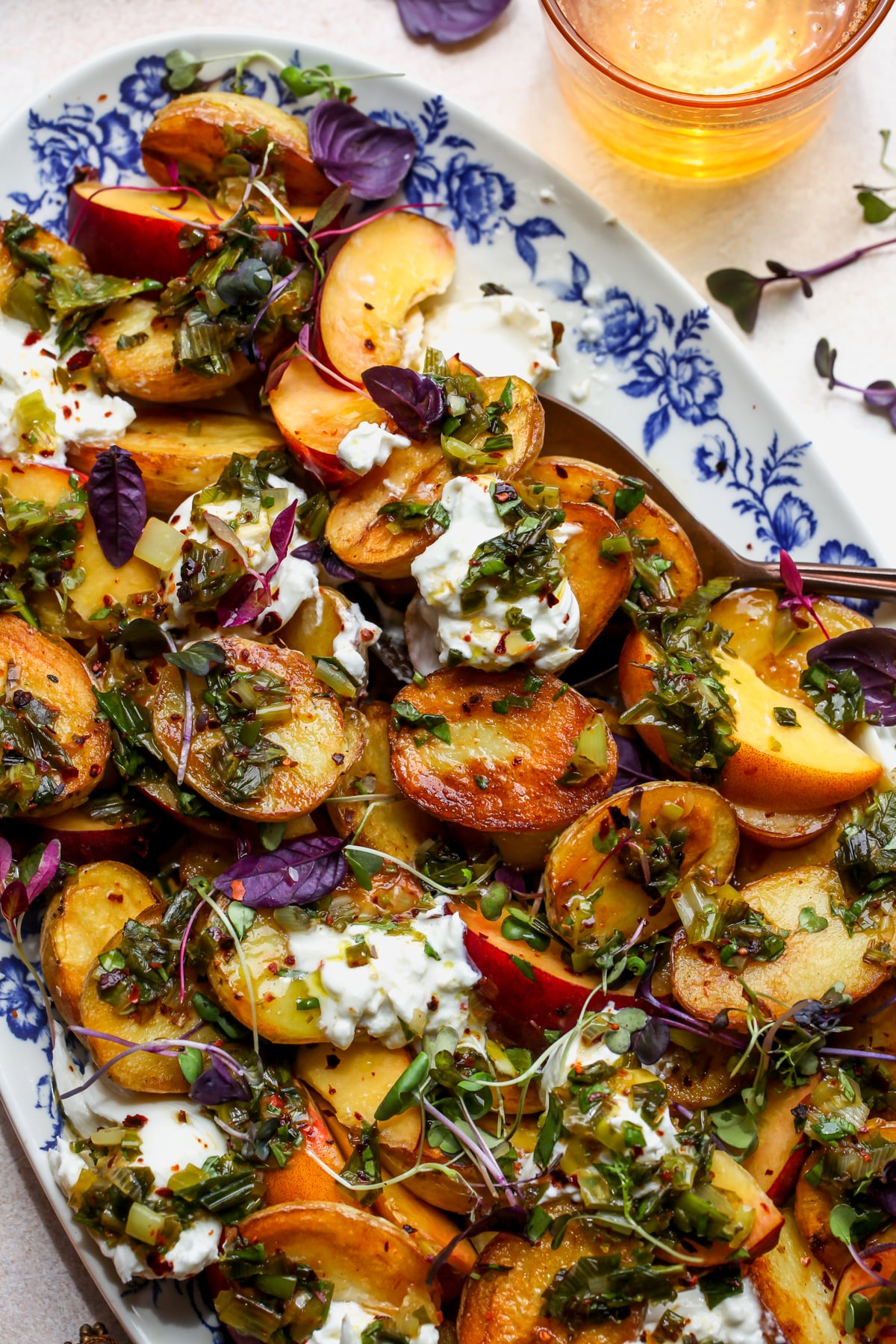 Crispy potato and peach salad with dollops of burrata and charred scallion dressing arranged on a white platter with blue floral design.