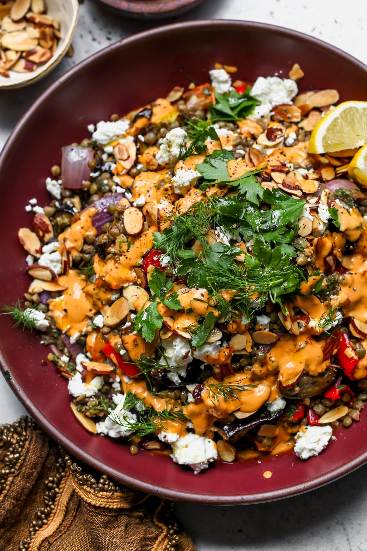 Lemony lentil and roasted eggplant salad drizzled with harissa-tahini and garnished with fresh herbs styled in a maroon serving bowl.