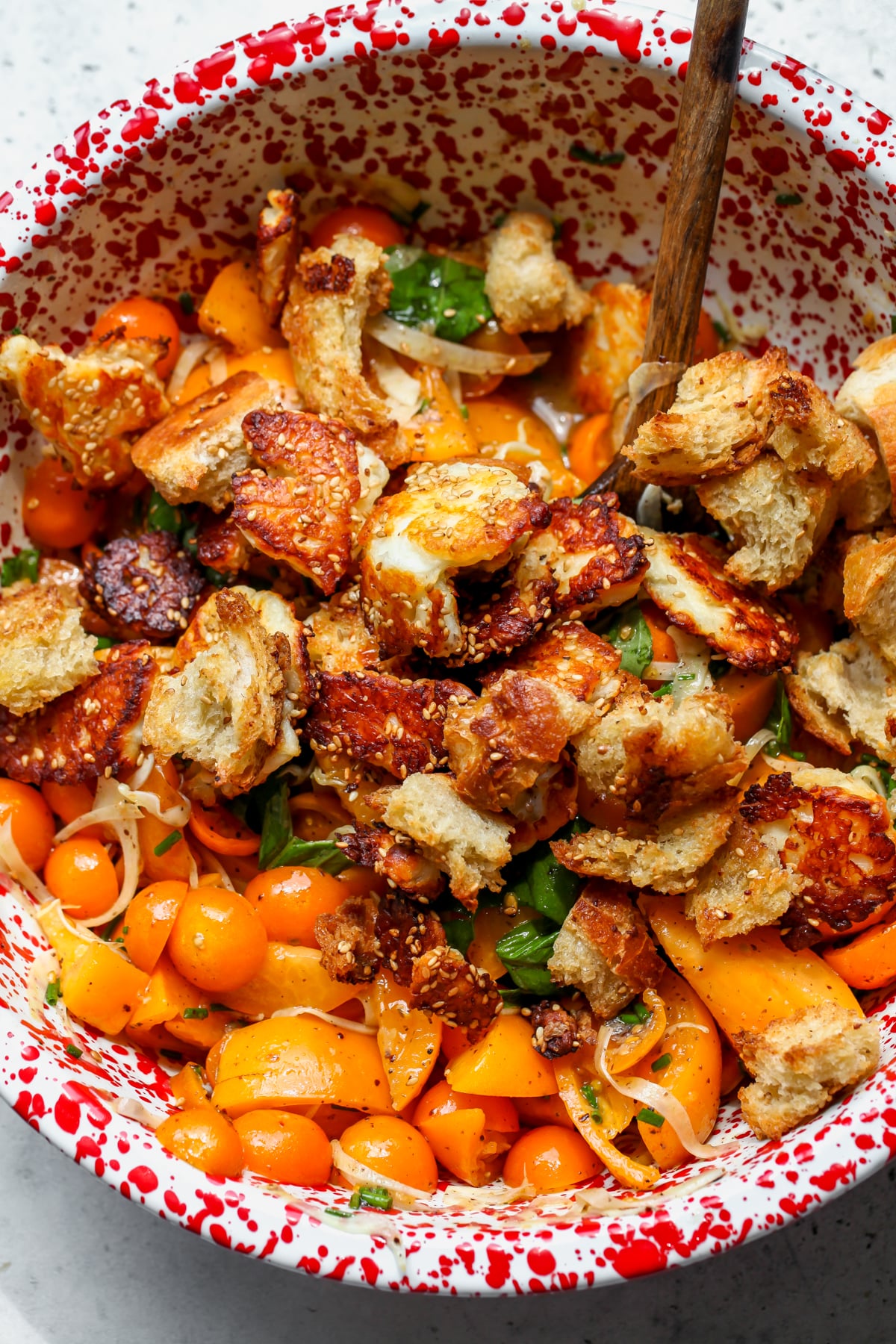 Baked halloumi and bread being tossed with tomato mixture in a large red speckled mixing bowl.