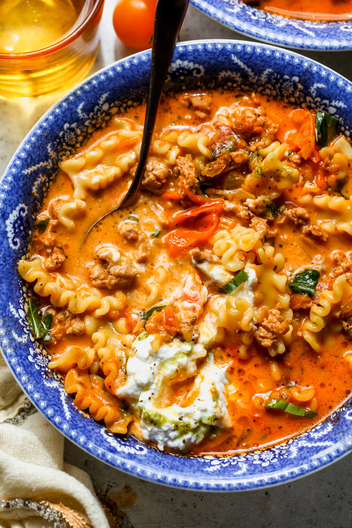 Lasagna soup topped with ricotta and pesto oil being stirred in a small blue soup bowl.