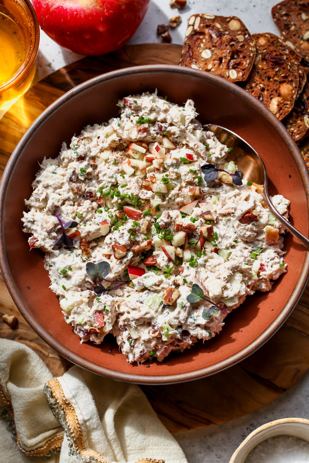 Apple-Chive Tuna Salad in a maroon bowl garnished with fresh shives and diced apple. Crackers arranged outside of the bowl.