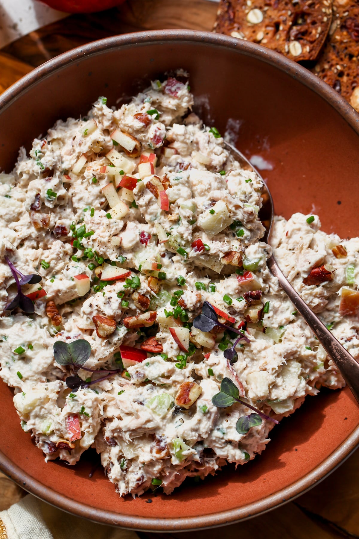 A spoon scooping up a portion of apple tuna salad in a maroon bowl.