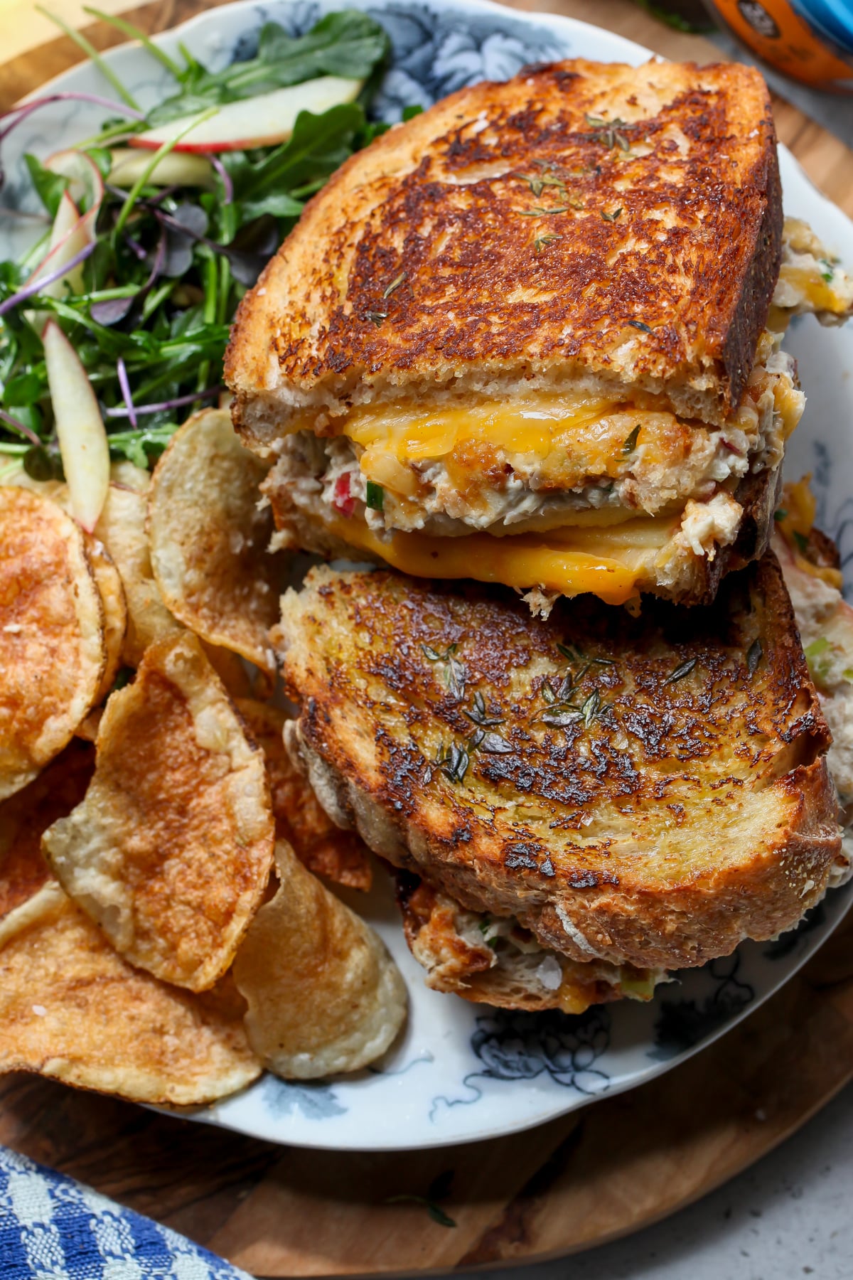 A tuna melt sliced in half and arranged on a white and blue floral plate with chips and arugula salad on the side.