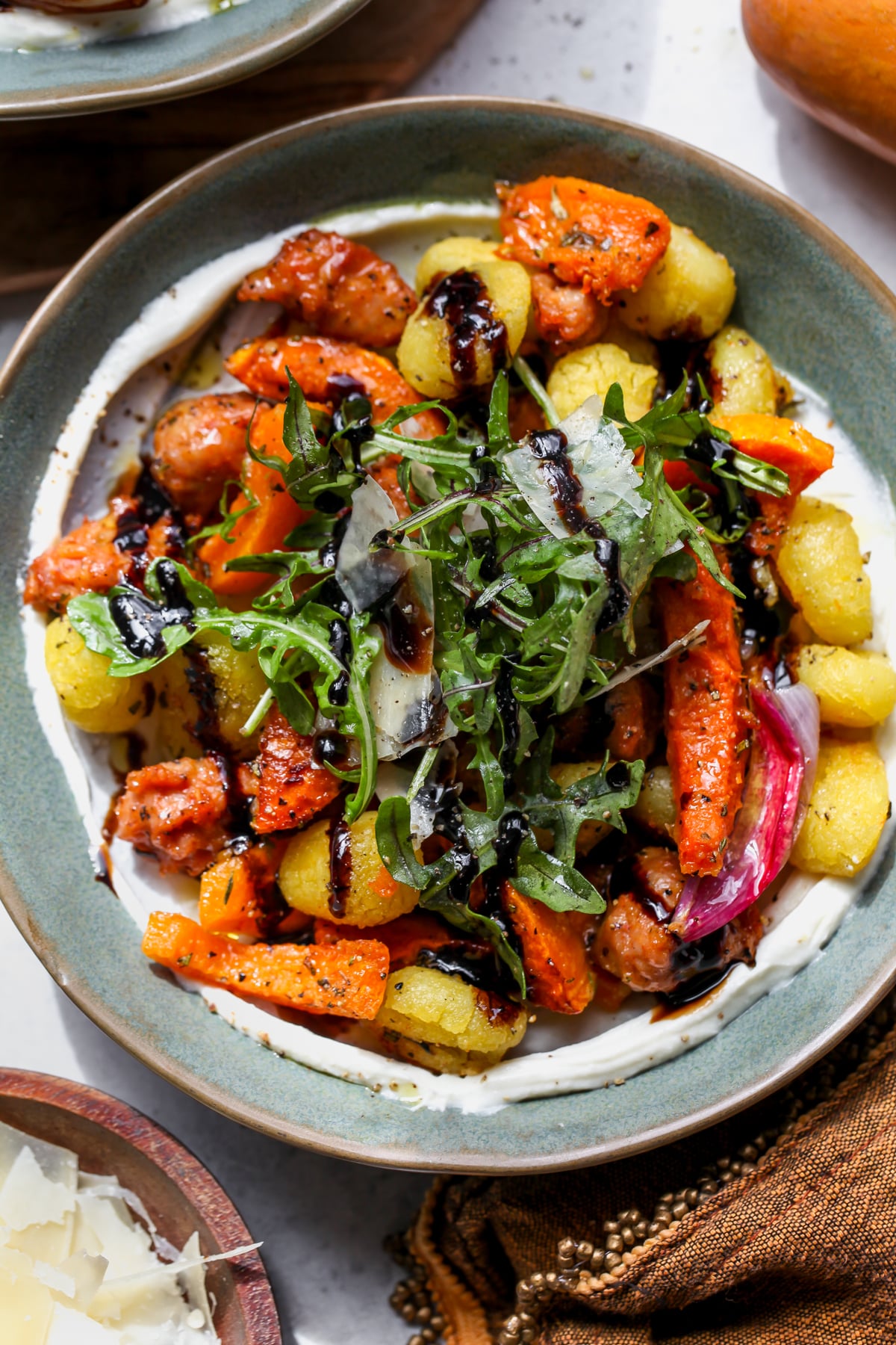 Whipped ricotta spread in a blue bowl topped with gnocchi, sausage, and squash, and garnished with arugula salad and balsamic glaze.